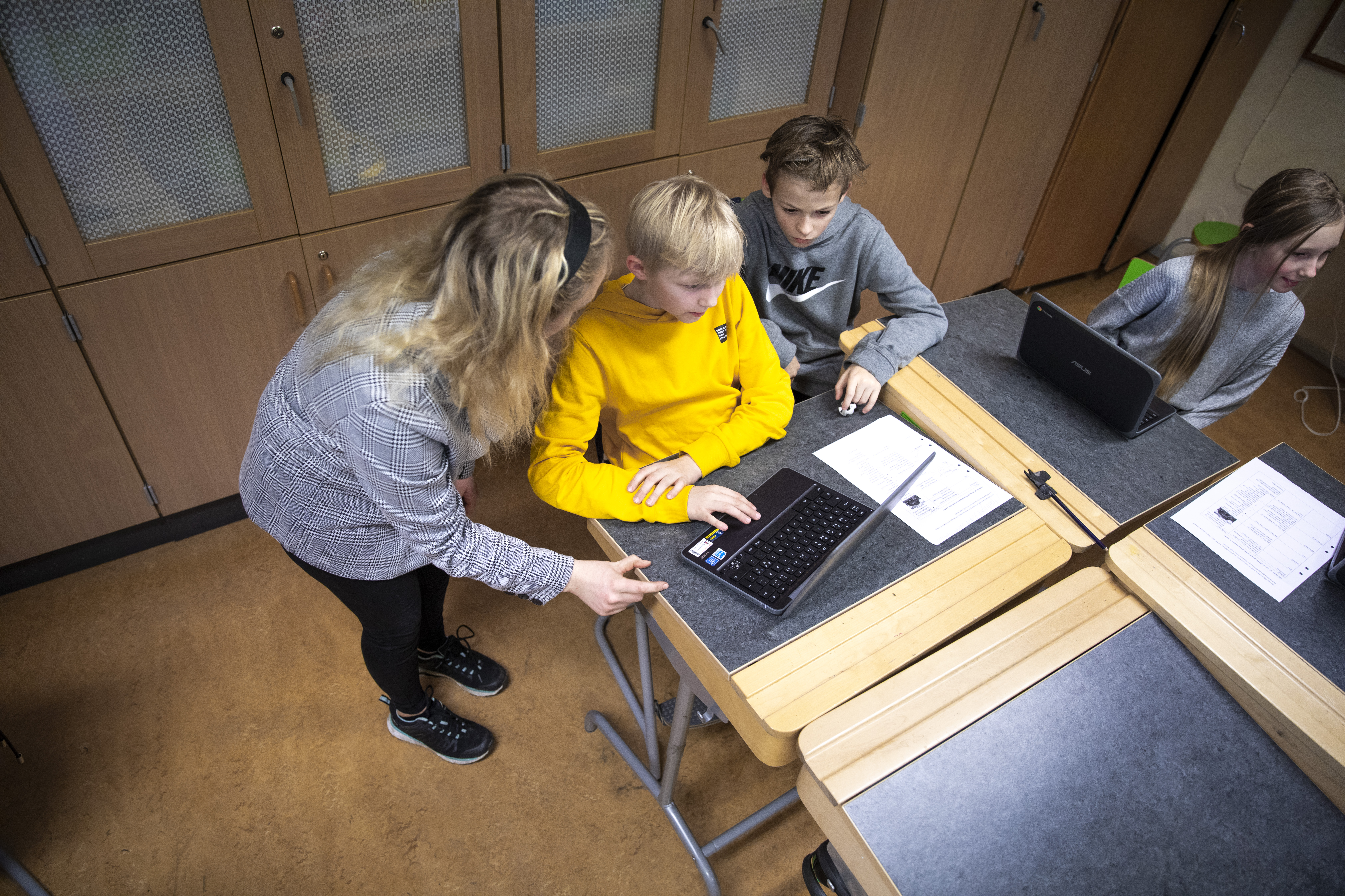 Lärare står i ett klassrum och hjälper två elever som sitter framför en dator. 